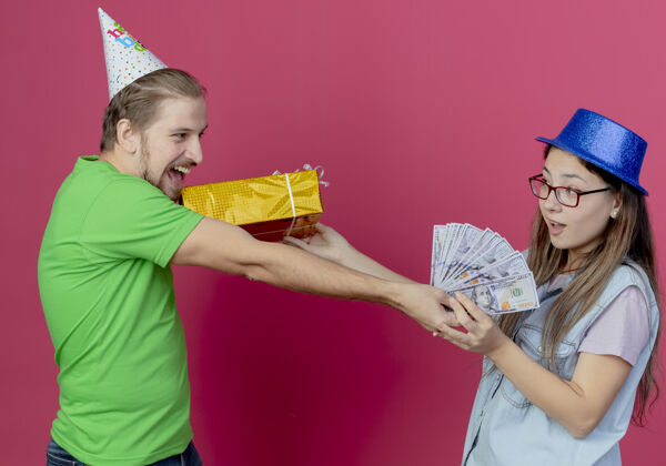 情侣一对戴着派对礼帽的年轻夫妇相互看着对方 手里拿着礼盒和钱 隔离在粉红色的墙上欢乐穿着礼物