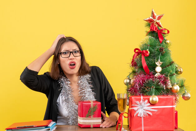 坐一位穿着西装 戴着眼镜 拿着礼物 坐在办公室里一棵圣诞树旁的困惑的商务女士时尚头发肖像