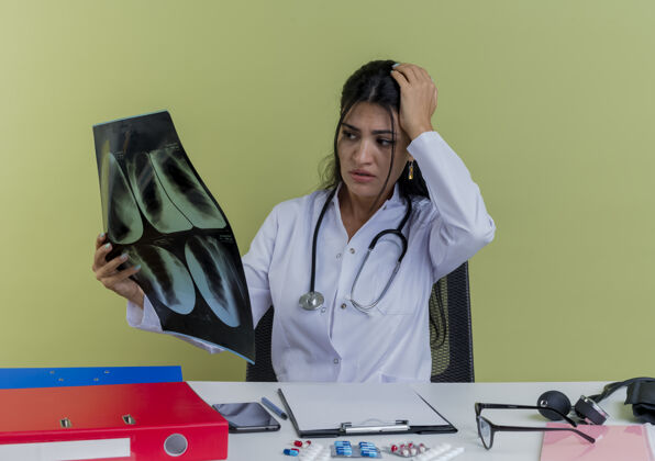 成人焦虑的年轻女医生穿着医用长袍和听诊器坐在办公桌旁 手里拿着医疗工具 看着x光片 手放在头上与世隔绝绿色穿保持