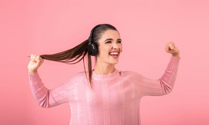 时髦年轻迷人的女人戴着无线耳机听音乐穿着粉色毛衣微笑快乐积极的心情摆在粉色背景上孤立搞笑的情绪表情微笑漂亮人