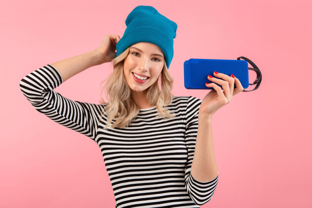 情感年轻漂亮的女人拿着无线扬声器听音乐穿着条纹衬衫和蓝色帽子微笑快乐积极的心情摆在粉红色的背景上舒适女性服装