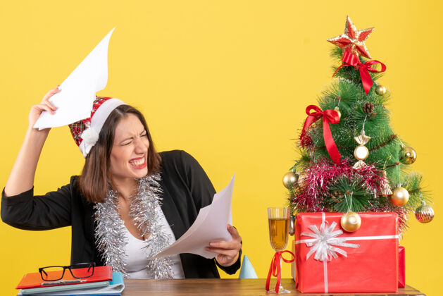 西装情绪化的商务女士穿着西装 戴着圣诞老人帽和新年装饰品 手里拿着文件 坐在一张桌子旁 桌子上放着圣诞树人快乐桌子