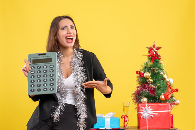 头发圣诞气氛 微笑的美丽女士站在办公室 指着黄色的办公室计算器黄色计算器女士