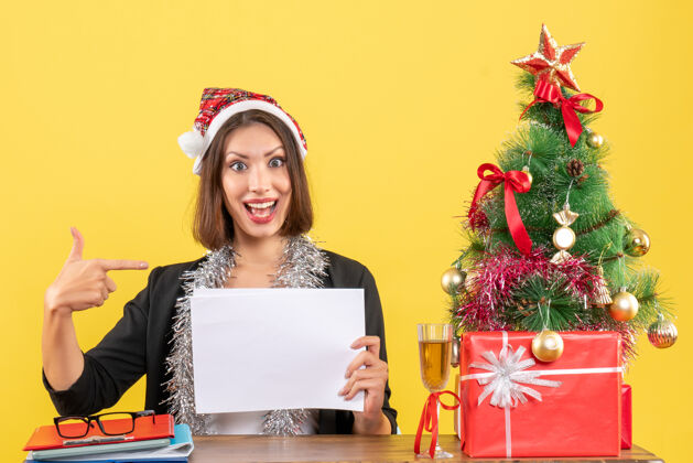 指着一位身着西装 头戴圣诞老人帽 戴着新年装饰品 面带微笑的商务女士独自一人指着文件 坐在办公室里一棵圣诞树上的桌子旁文件帽子坐着