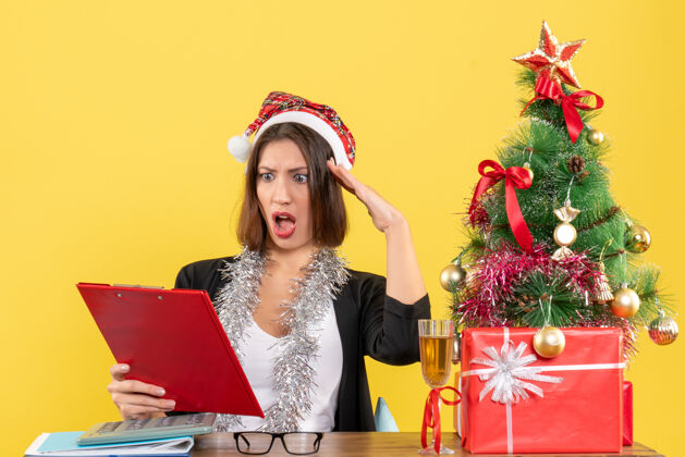 人穿着西装 戴着圣诞老人帽 戴着新年装饰品的商务女士看着文件 坐在一张桌子旁 桌子上放着圣诞树坐树圣诞老人
