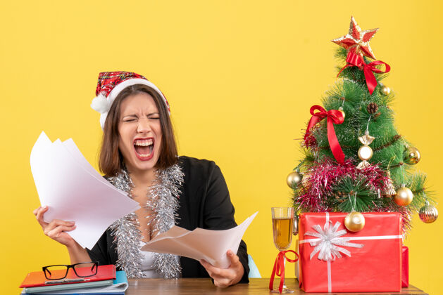 性感情绪紧张的商务女士穿着西装 戴着圣诞老人帽和新年装饰品 手里拿着文件 坐在一张桌子旁 桌子上放着圣诞树情绪圣诞老人桌子