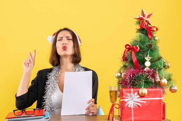 帽子一位穿着西装 戴着圣诞老人帽 戴着新年装饰品的梦幻商务女士独自一人指着头顶 坐在办公室里一张桌子旁 桌子上放着圣诞树人树年