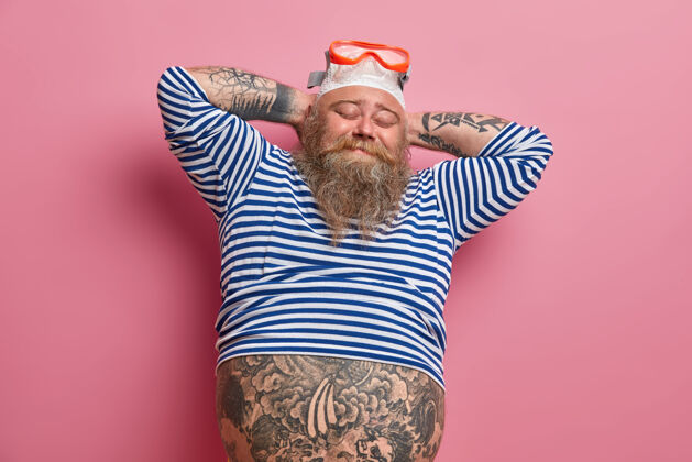 庆祝放松的纹身男游泳者双手放在脑后 闭上眼睛 戴着泳镜和泳帽 大肚子从粉红色墙上的小水手衬衫上露出来暑假游泳运动员肥胖胡子