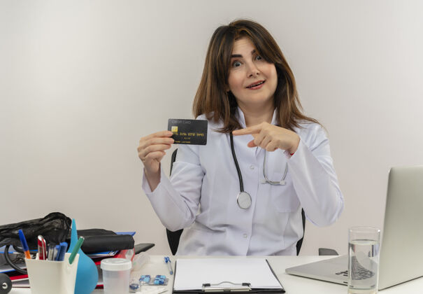 女性印象深刻的中年女医生穿着医用长袍和听诊器坐在办公桌前 拿着医疗工具剪贴板和笔记本电脑拿着 指着信用卡持有印象女士