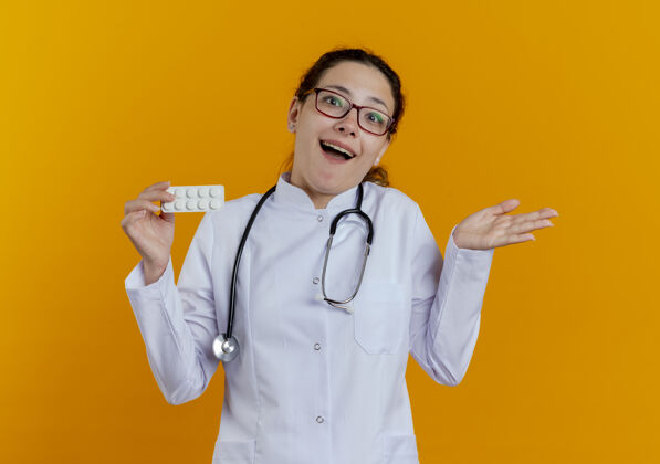 听诊器快乐的年轻女医生 穿着医用长袍 戴着听诊器 戴着眼镜 拿着药片 双手分开成人人女人
