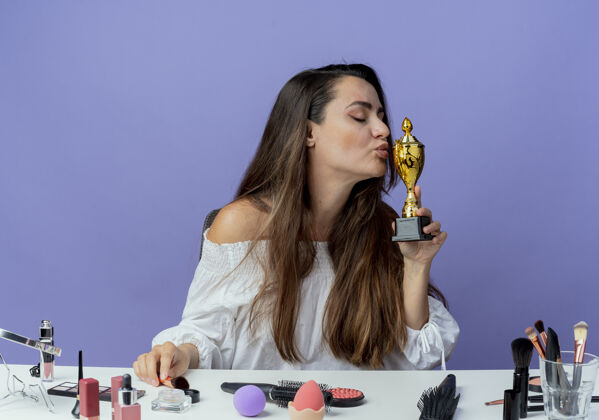 女人高兴的漂亮女孩坐在桌旁用化妆工具假装亲吻隔离在紫色墙上的优胜者杯工具化妆品优胜者