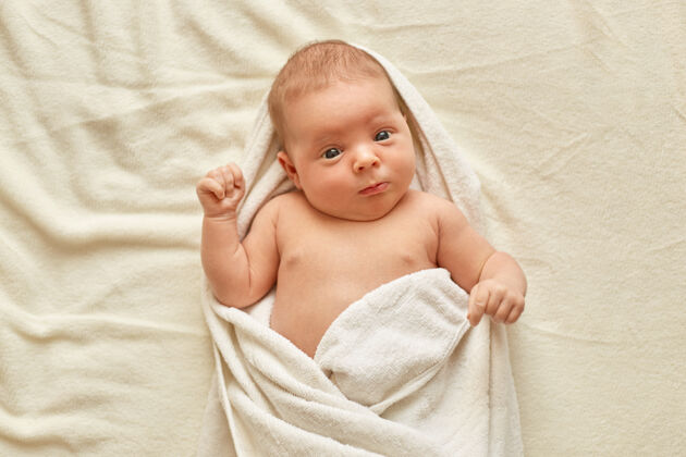 浴室刚出生的宝宝洗澡后穿着毛巾 可爱的小宝宝躺在白毯子上的床上 看着和学习外面的东西童年和宝宝的护理家庭房子洗衣服