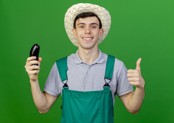 花园微笑着的年轻男园丁戴着园艺帽 拿着茄子 竖起大拇指 在绿色的背景上与复制空间隔离开来复制绿色年轻
