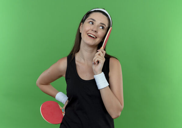 头带戴着头巾拿着球拍打乒乓球的年轻健身女士站在蓝色背景下 面带微笑地看着一边年轻微笑网球