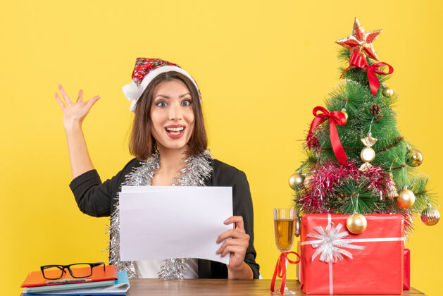 坐着穿着西装 戴着圣诞老人帽 戴着新年装饰品的商务女士独自一人指着身后拿着文件 坐在一张桌子旁 桌子上放着圣诞树帽子文件桌子