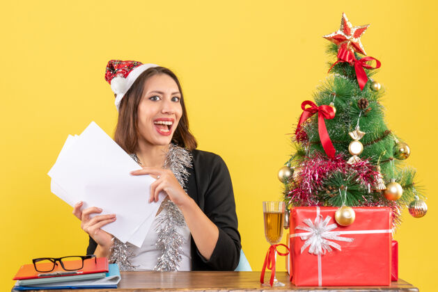 成人穿着西装 戴着圣诞老人帽和新年装饰品的商务女士独自工作 坐在办公室的一张桌子旁 桌上放着圣诞树快乐独自树