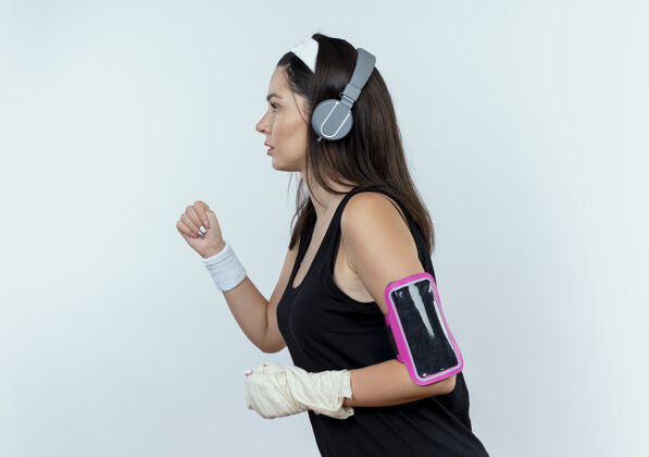 耳机戴着耳机和智能手机臂带的年轻健身女士站在白色背景下锻炼身体臂带手机站立