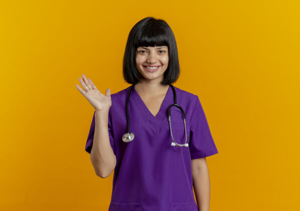 制服带着微笑的年轻黑发女医生穿着制服 手持听诊器站在橙色背景上 举手分开 留有复印空间年轻手黑发