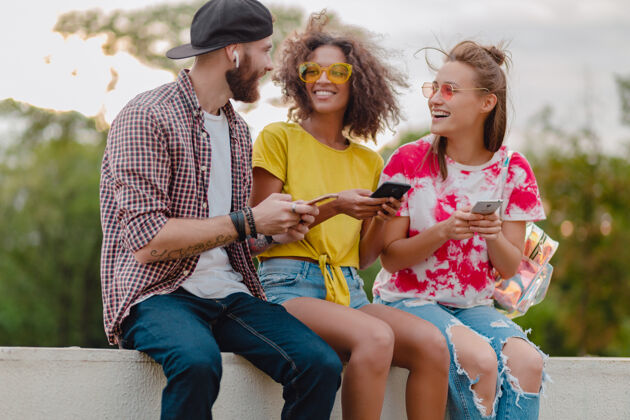 学生快乐的年轻朋友们坐在公园里用智能手机微笑 男人和女人玩得开心欢笑积极太阳镜