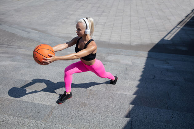 女性一个穿着衬衫 戴着白色耳机的年轻运动女性 在户外街头听音乐 带球做弓箭步 健康的生活方式 运动 活动 减肥的概念城市健身减肥