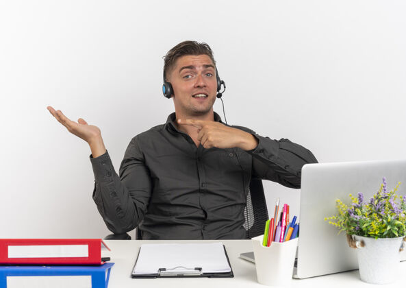 点年轻自信的金发上班族戴着耳机坐在办公桌旁 拿着办公工具 用笔记本电脑指着空手 在白色背景上隔离 留有复印空间年轻金发耳机