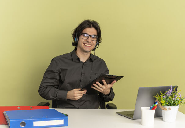 剪贴板戴着耳机 戴着眼镜 面带微笑的年轻上班族坐在办公桌旁 手里拿着办公工具 手里拿着笔记本电脑 把剪贴板隔离在绿色背景上 还有复印空间眼镜微笑持有