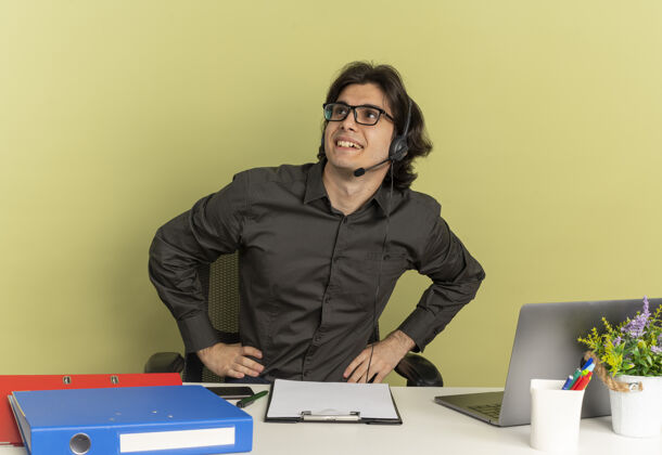 年轻戴着耳机 戴着眼镜 面带微笑的年轻上班族坐在办公桌旁 手里拿着办公工具 手里拿着笔记本电脑 看着绿色背景上隔离的一面 还有复印空间壁板办公室眼镜