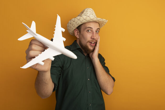 男人兴奋的金发帅哥 戴着沙滩帽 把飞机模型孤立地放在橙色的墙上帽子举行模特