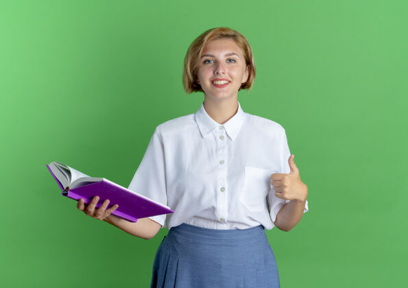 女孩年轻微笑的金发俄罗斯女孩竖起大拇指 拿着一本书 背景是绿色的 还有复印空间微笑拇指抱着