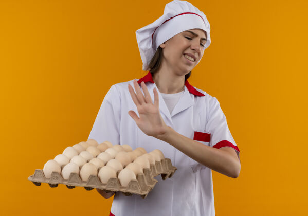 年轻穿着厨师制服的年轻恼怒的白人厨师女孩手持并假装用手推着一批鸡蛋 这些鸡蛋被隔离在橙色背景上 留有复制空间假装批举行