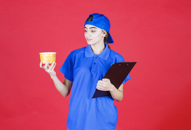 智能穿着蓝色制服的女快递员手里拿着一个黄色的面杯和一个黑色的文件夹年轻人呆在家里年轻人
