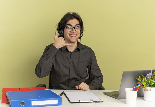 坐着戴着耳机 戴着眼镜 面带微笑的年轻上班族坐在办公桌旁 手里拿着办公工具 手里拿着笔记本电脑的手势 手机手势被隔离在绿色背景上 还有复印空间耳机光学手势