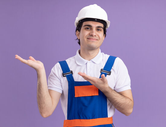 建设者身穿施工制服 头戴安全帽的年轻建筑工人站在紫色的墙壁上 双臂欢快地微笑着展示复制品空间微笑年轻人站立