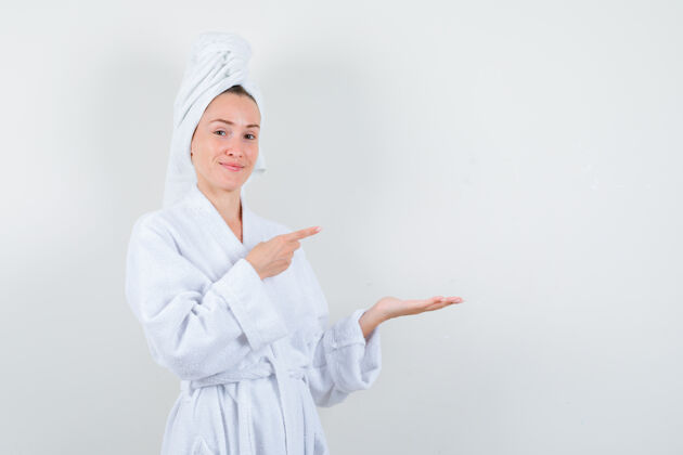 乐趣穿着白色浴袍的年轻女子 手巾指着她摊开的手掌 看上去很愉快 正对着前方指向模特头发