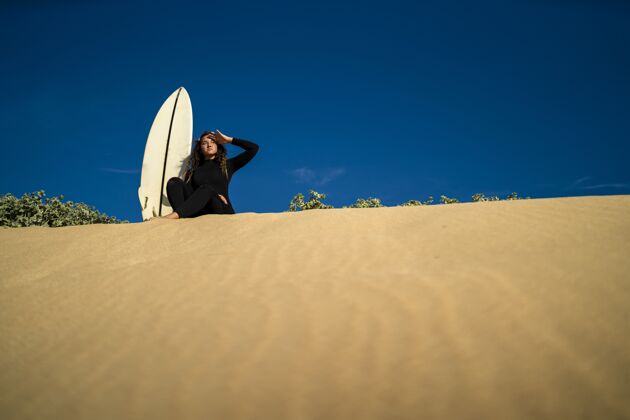 阳光低角度拍摄的一个迷人的女性坐在沙山与冲浪板在一边人旅游表面