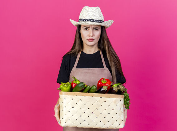 园艺穿着制服的漂亮的园丁女孩戴着园艺帽 手里拿着粉红色背景上孤立的菜篮子篮子持有帽子