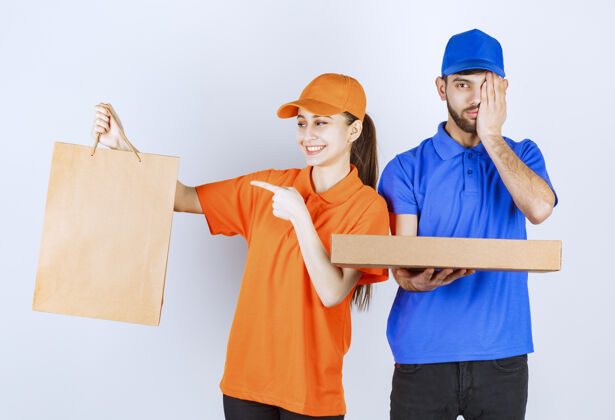 模特穿着蓝黄制服的快递员男孩和女孩拿着纸板外卖盒和购物包 看上去既困惑又害怕压力食物订单