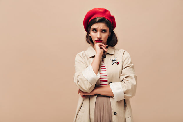 成人穿着时髦战壕和贝雷帽的悲伤女人在米色背景上摆姿势一个戴着红帽子 穿着条纹毛衣和雨衣的女孩在镜头前摆姿势巴黎姿势黑发