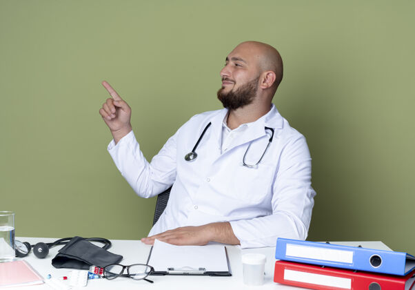 请穿高兴的年轻秃头男医生穿着医用长袍和听诊器坐在办公桌前 用医用工具点在一旁 隔离在绿色背景上秃头医疗坐着