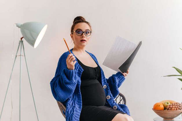 女性戴眼镜的孕妇有主意穿着深蓝色开衫和连衣裙的迷人女士拿着笔记本和笔坐在木椅上孕妇肚子成人