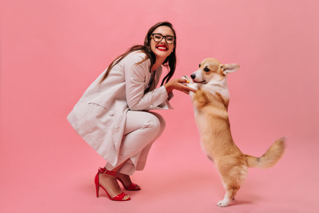 休闲戴着眼镜和西装的女士在粉色背景下和高尔基犬玩耍穿着办公室服装和红色高跟鞋的快乐女人微笑着抱着高尔基犬时尚狗空白