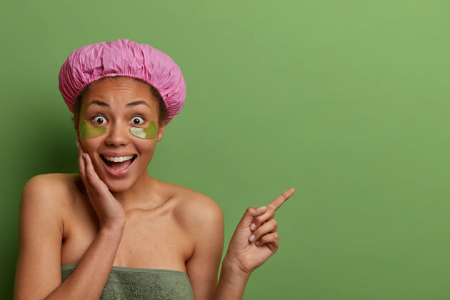 微笑惊喜快乐的黑皮女人在眼部应用抗衰老胶原蛋白贴片 显示出奇的要价 点在空格上 隔离在绿色墙壁上 戴浴帽 毛巾绕身体成人满意一旁