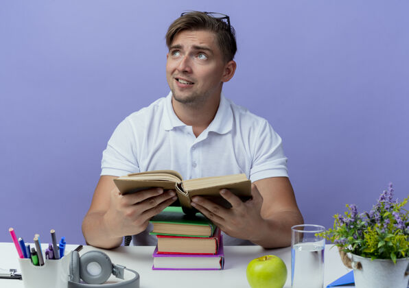 侧线看着旁边高兴的年轻英俊的男学生坐在书桌旁 手里拿着书本 背景是蓝色的请抱着书桌
