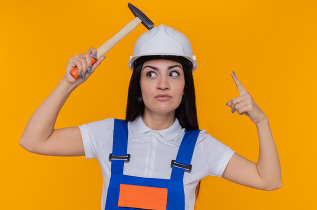 表演身穿施工制服 头戴安全帽的年轻建筑工人站在橙色的墙上 头上举着锤子 食指微笑着 有着伟大的想法锤子头盔手指