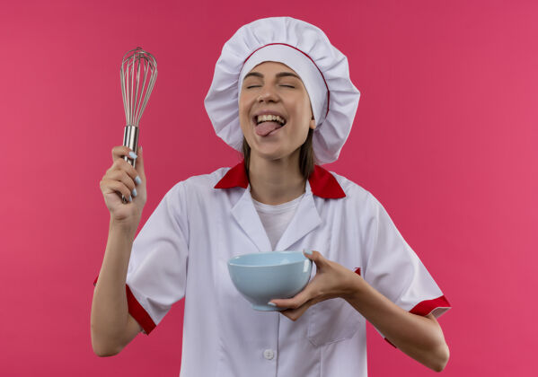 烹饪穿着厨师制服的年轻快乐的白人厨师女孩拿着打蛋器和碗 把舌头伸到粉红色的地板上 留着复印空间厨师复制粉红
