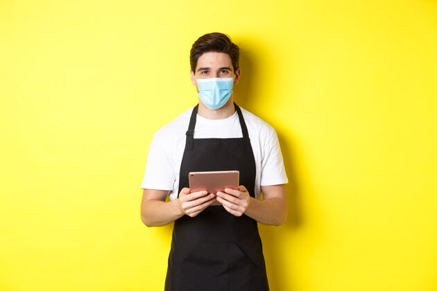 面膜covid-19的概念 小型企业和流行病穿着黑色围裙和医用面罩的服务员拿着数字平板电脑 站在黄色背景上男士男士成人