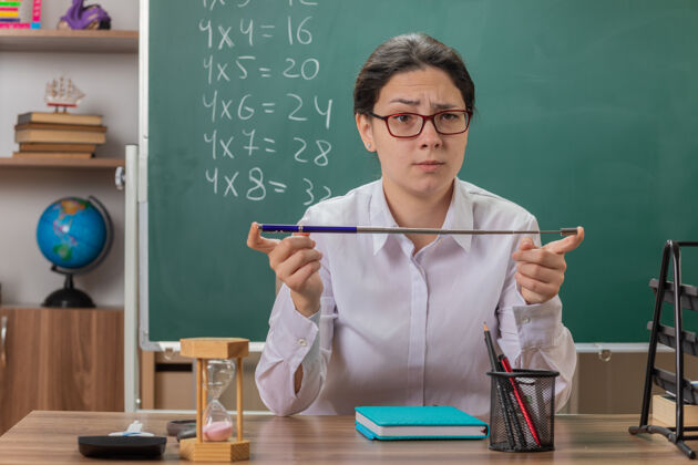 戴着年轻的女老师戴着眼镜 严肃地看着前面 拿着指针 坐在教室黑板前的课桌上讲解课文女人认真年轻人