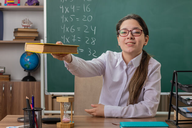 奉献年轻的女教师戴着眼镜 面带微笑 自信地坐在教室黑板前的课桌旁看书自信穿着女人