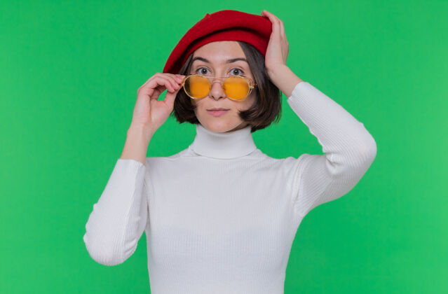 立场戴着贝雷帽 戴着黄色太阳镜 留着短发 穿着白色高领毛衣的年轻女子站在绿色迷茫的墙前看着前方太阳镜短裤贝雷帽