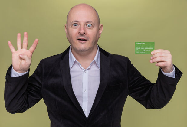 微笑一个穿着西装的中年秃头男人拿着信用卡 面带微笑 惊讶地看着前面的四号 手指站在绿色的墙上四手指秀